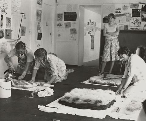A black-and-white photograph of four women participating in an indoor creative workshop, working on felt pieces. The walls are covered in drawings and notes.